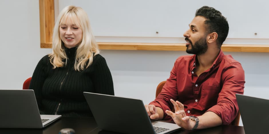 Two Awin employees sitting at a desk in a meeting room, engaging in a discussion while using their laptops.